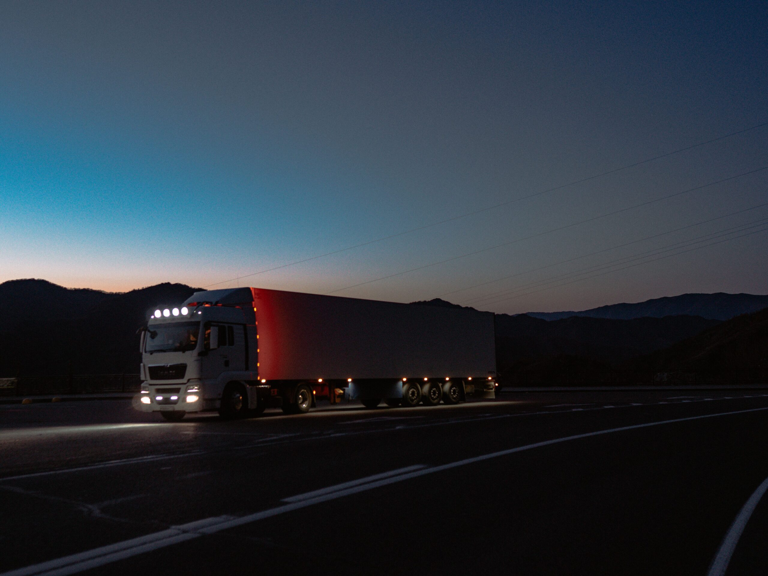 A semi truck driving on a road at dusk, with a colorful sky and headlights illuminating the path ahead.